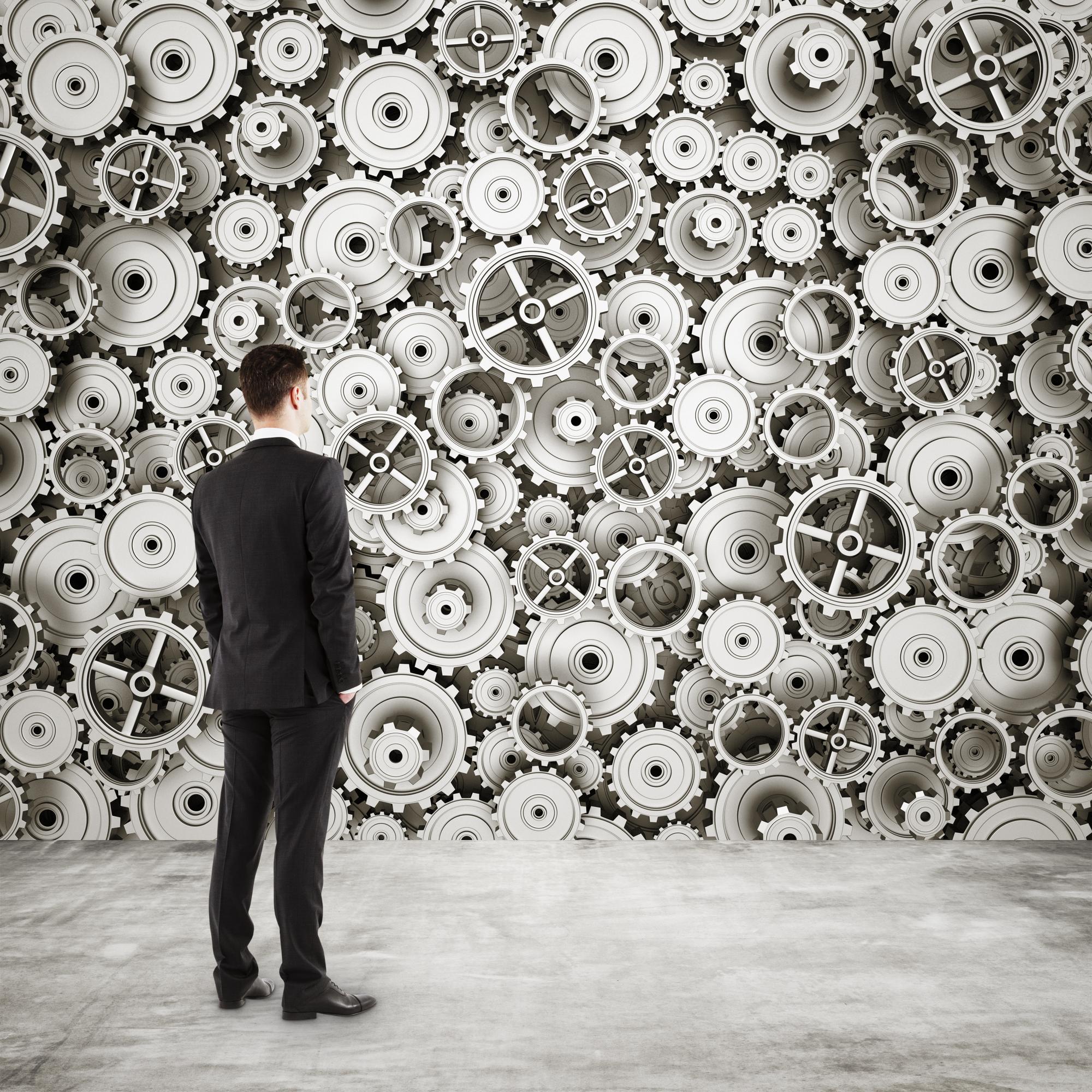 تصویر فردی که به دیواری پر از چرخ‌دنده‌ها و سیستم‌های پیچیده نگاه می‌کند، نمادی از تفکر استراتژیک و بهبود عملکرد.